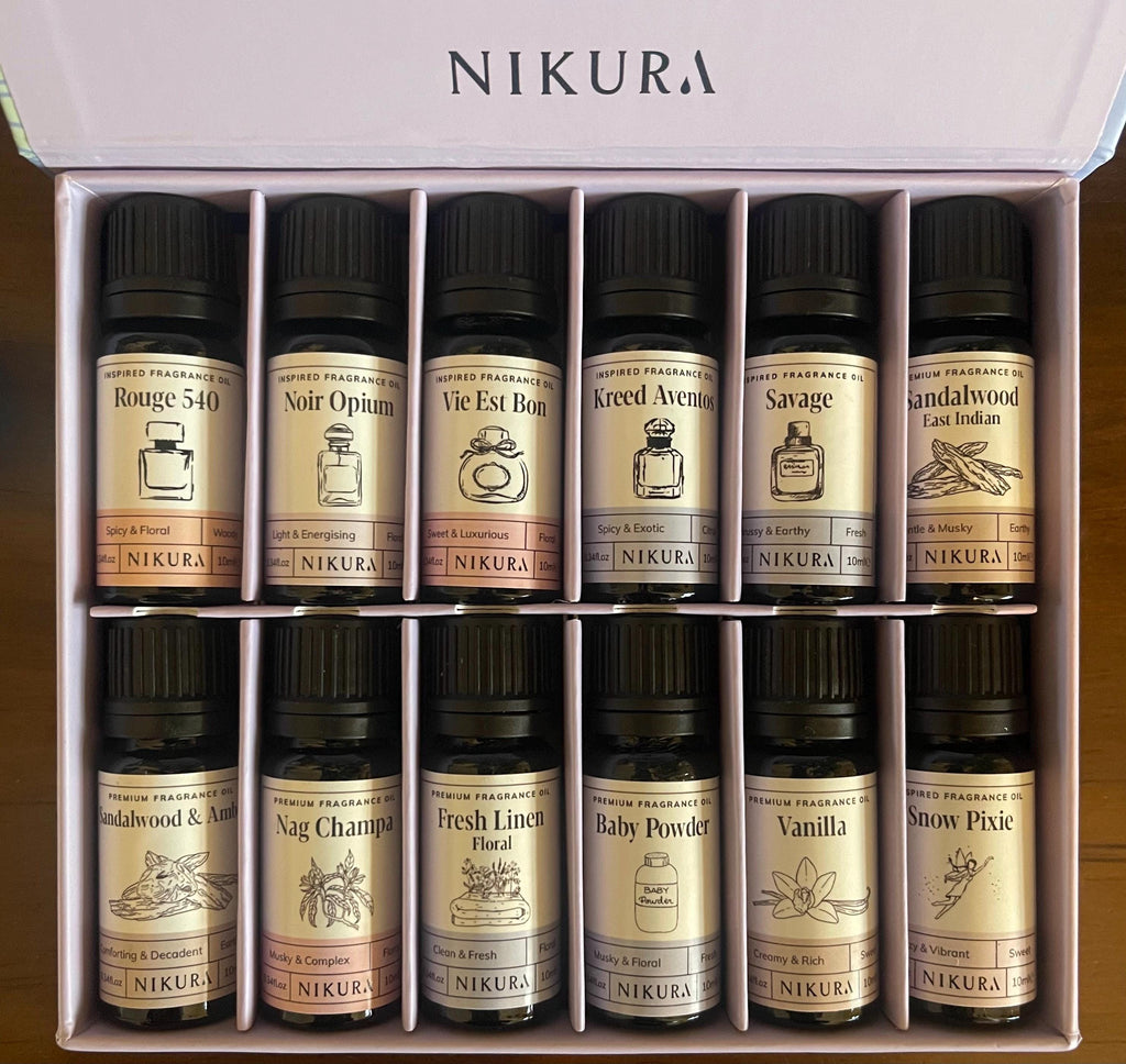 A boxed set of 15 Nikura essential oils arranged in a 3x5 grid inside an open white presentation box. The set includes scents such as Bourge 540, Noir Opium, Vie Est Bon, Acreed Aventure, Savage, Landalwood, Naje Champi, Fresh Linen, Baby Powder, Vanilla, and Snow Pixie.