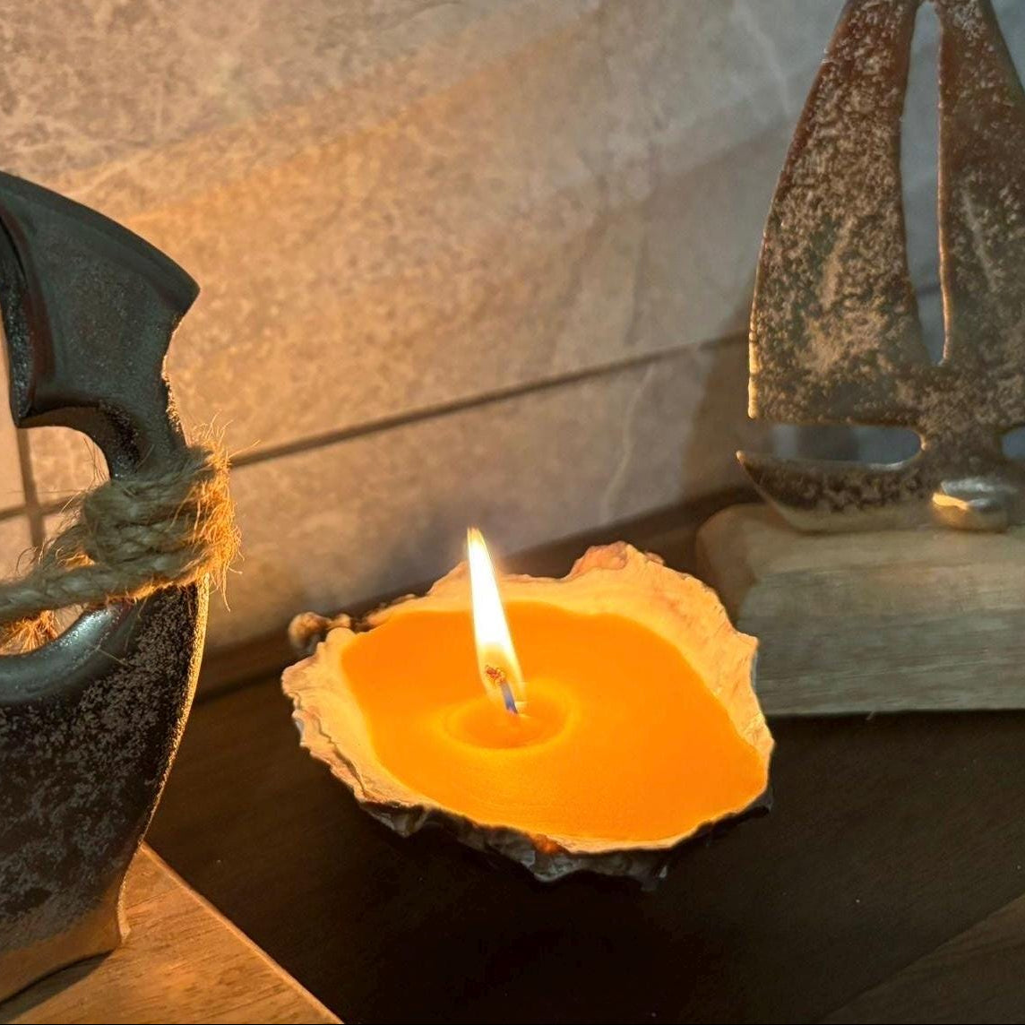 A lit orange candle in a rustic shell holder sits on a dark wooden surface, flanked by decorative nautical sculptures against a tiled wall.
