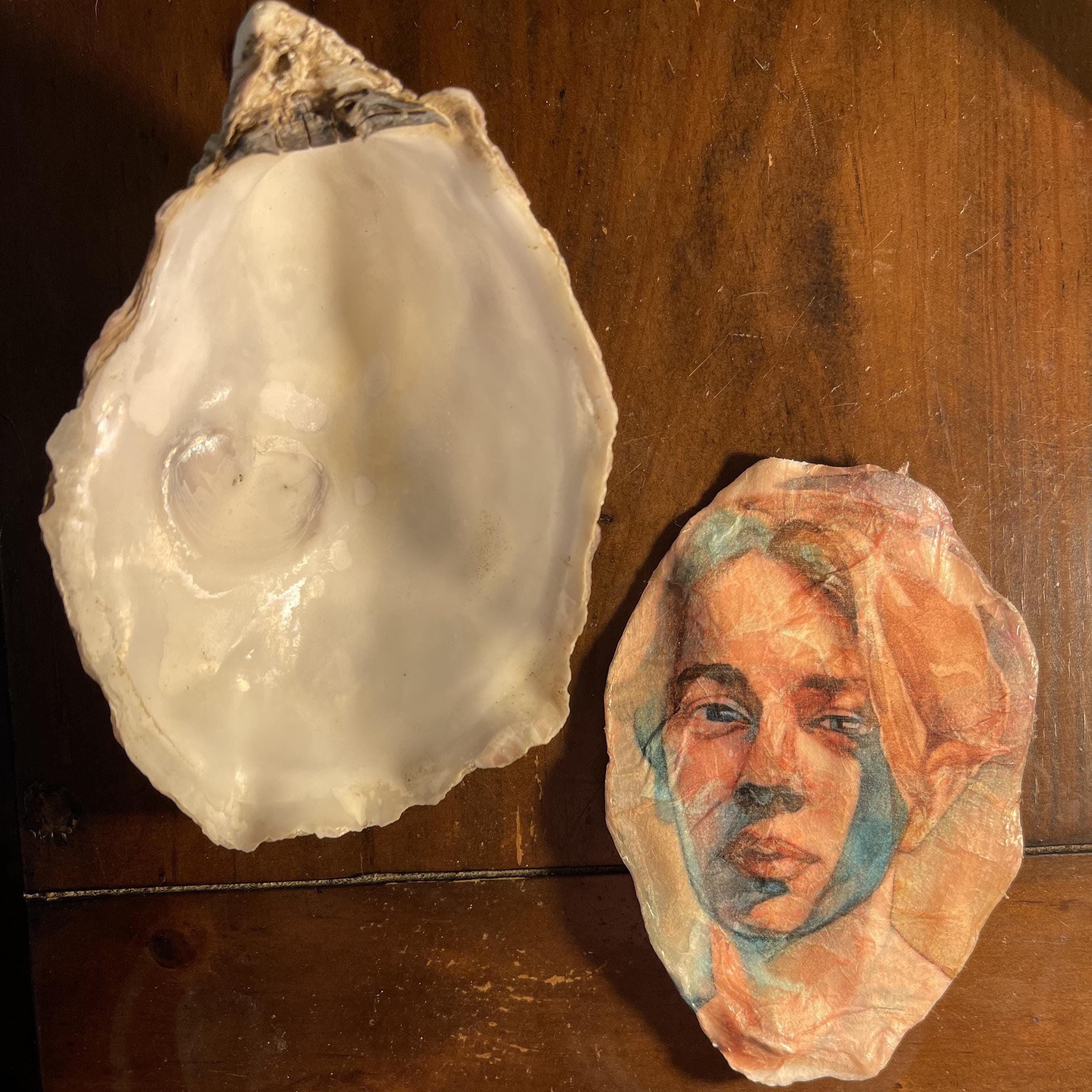 Two oyster shells displayed on a wooden surface, with one shell showing a glossy white interior and the other featuring a painted portrait of Tom Odel, a musician in soft peach, blue, and brown tones.