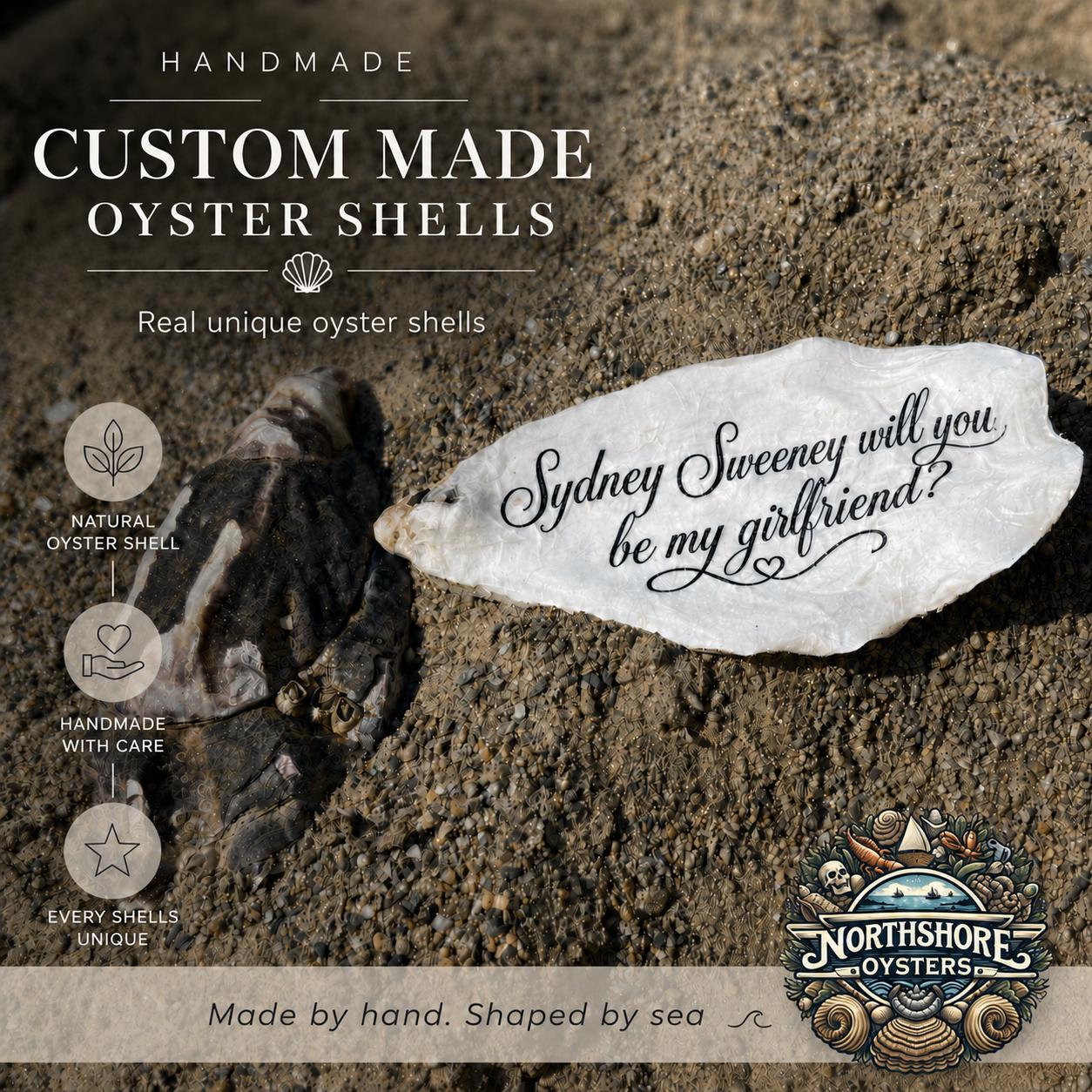 A custom engraved oyster shell with the phrase 'will you be my girlfriend?' displayed on a sandy beach next to a natural dark oyster shell. The image features promotional text highlighting handmade and custom-made oyster shells.
